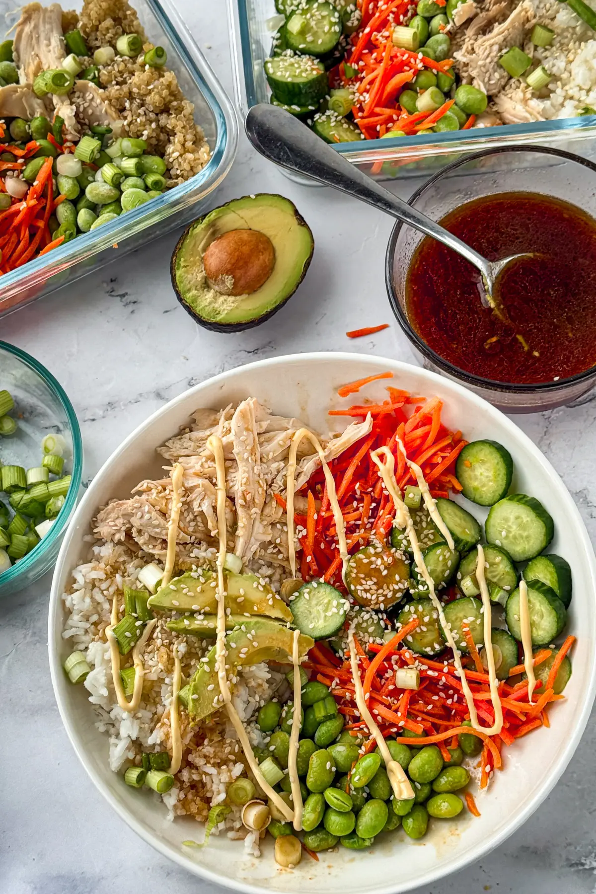 An Asian-inspired chicken and rice power bowl on a table next to a bowl of sauce and two meal prep containers (already prepped).
