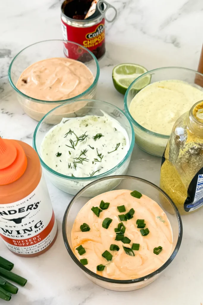 The sauces in glass bowls spread out on a counter next to the ingredients, like a bottle of Kinder's and a bottle of Dijon mustard.