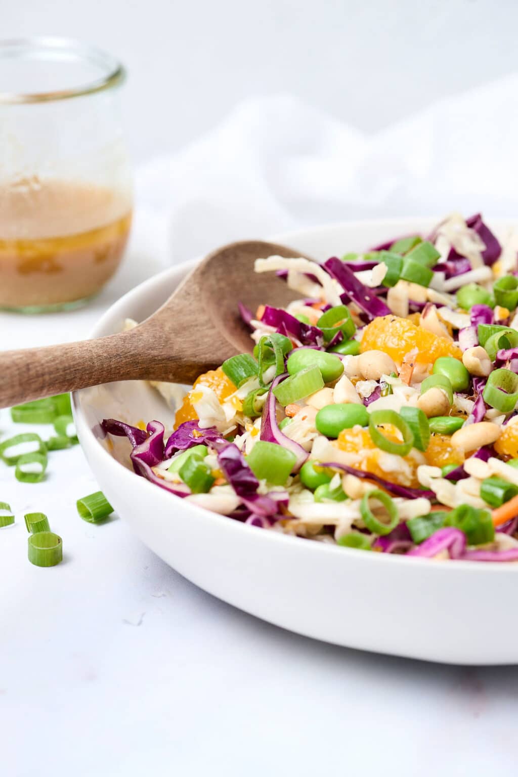 A close-up of an Asian cabbage salad with edamame in a bowl with a wodden tong in it on a table next to dressing.
