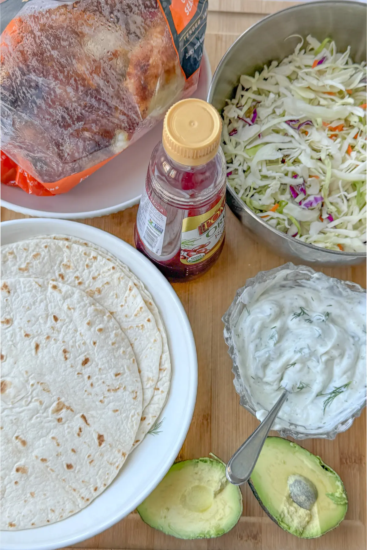 A rotisserie chicken, bowl of coleslaw, plate of wraps, Greek yogurt ranch, a bottle of red wine vinegar, and an avocado cut in half on a wooden board.
