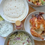 A birds-eye view of a rotisserie chicken, tortillas, and coleslaw on a board next to a large bowl of caprese pasta salad.