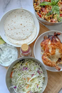 A birds-eye view of a rotisserie chicken, tortillas, and coleslaw on a board next to a large bowl of caprese pasta salad.