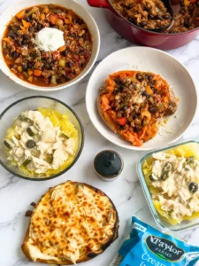 Multiple different meals on a counter: a pot of chili, a bowl of chili, a sweet potato stuffed chili, jalapeno popper shredded chicken over spaghetti squash, and a chicken flatbread next to a bagged salad.