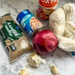 A carton of rolled oats, a bag of lentils, a can of white beans, garlic cloves, an onion, and a freezer bag of frozen rice on a counter.