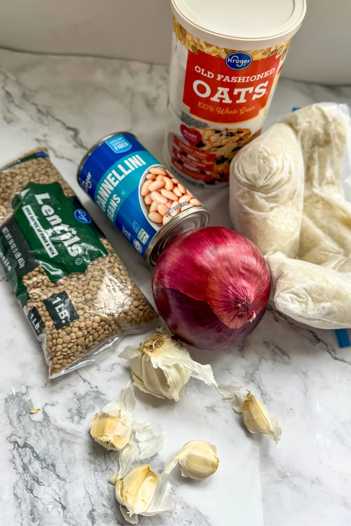 A carton of rolled oats, a bag of lentils, a can of white beans, garlic cloves, an onion, and a freezer bag of frozen rice on a counter.