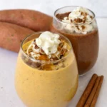 Two sweet potato smoothies topped with whipped cream on a counter next to sweet potatoes.