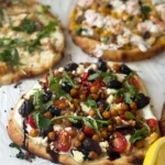 A Mediterranean veggie flatbared, a cottage cheese hot honey flatbread and street corn chicken flatbreads on parchment paper next to a lemon.