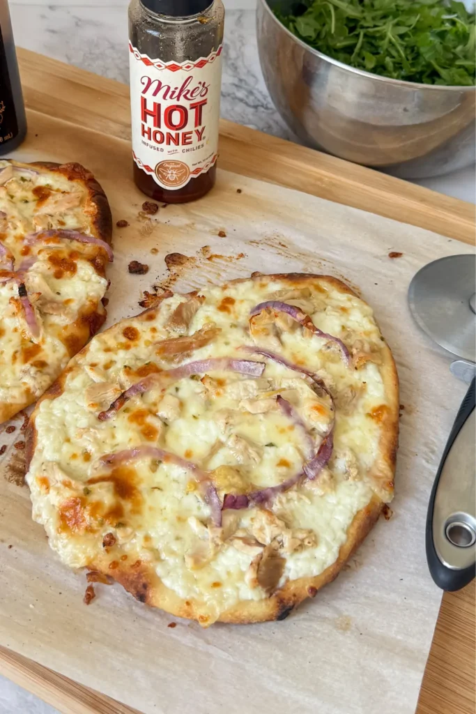 A cottage cheese flatbread on parchment paper on a wooden cutting board next to a bottle of hot honey.