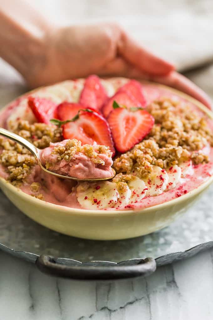 A strawberry banana smoothie bowl topped with homeamde granola and strawberries on a plate with a spoon in it.