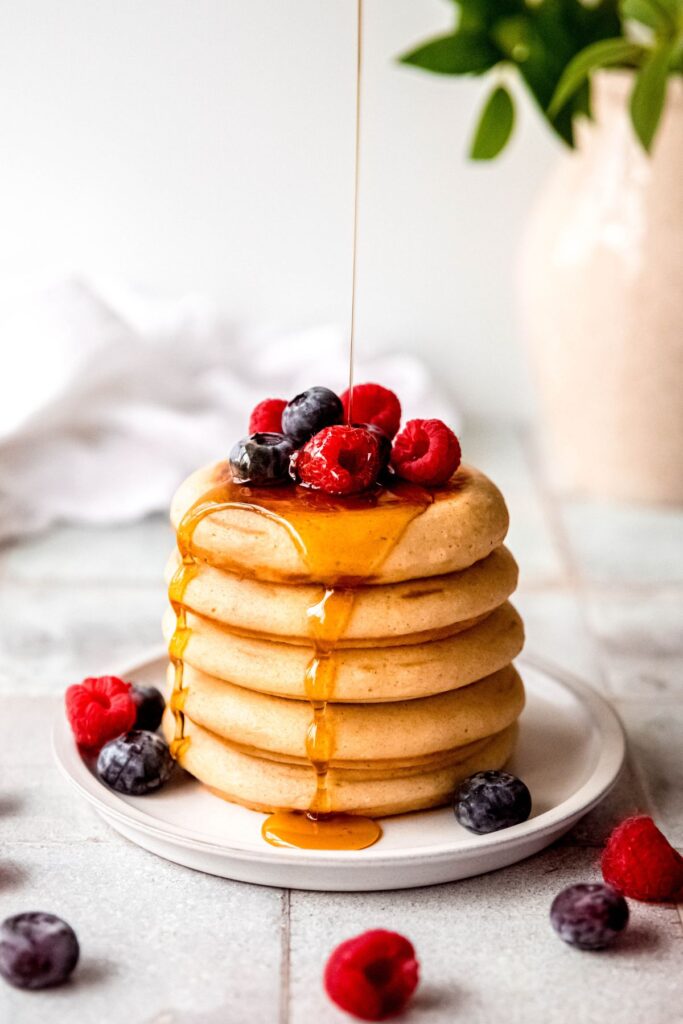 A stack of whole wheat pancakes with protein powder on a plate topped with berries and drizzle with syrup on a table. 