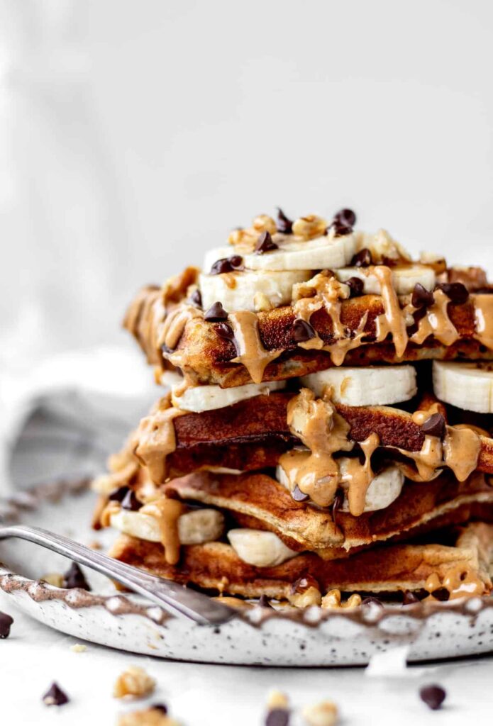 A stack of protein waffles on top of one another with peanut butter drizzling down the sides and banana slices on top.