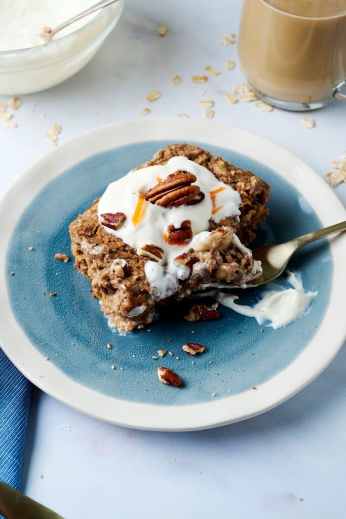 A slice of carrot cake baked oatmeal topped with a cream cheese and yogurt topping on a plate with a fork. 