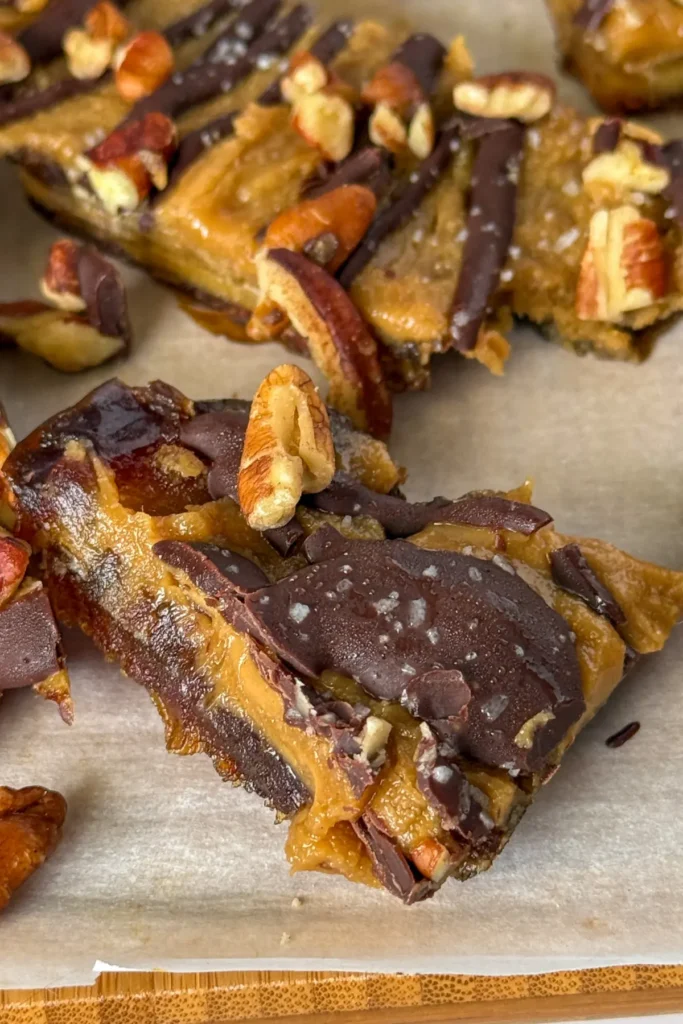 A close-up of a maple pecan bar drizzled in dark chocolate on parchment paper on a wood cutting board.