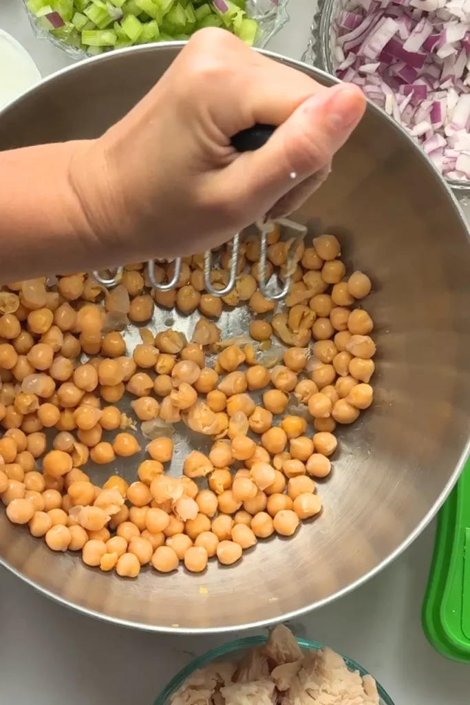 A potato masher mashing chickpeas in a mixing bowl on a counter.