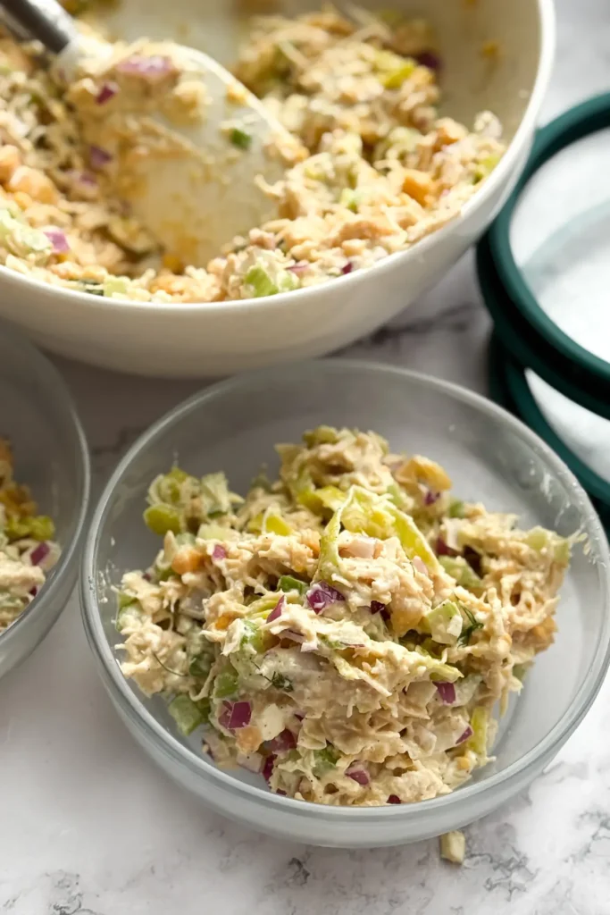 Portioned out chicken salad in a meal prep container next to a large bowl of pepperoncini salad.