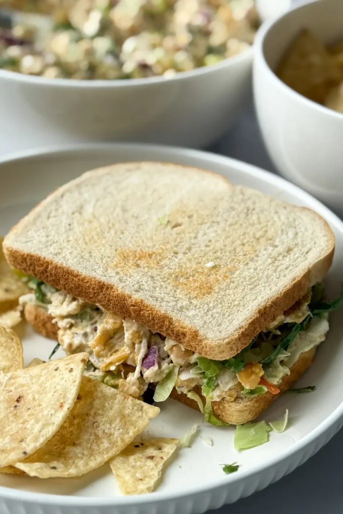A close-up of a chickpea and chicken salad sandwich on a plate next to tortilla chips on a table.