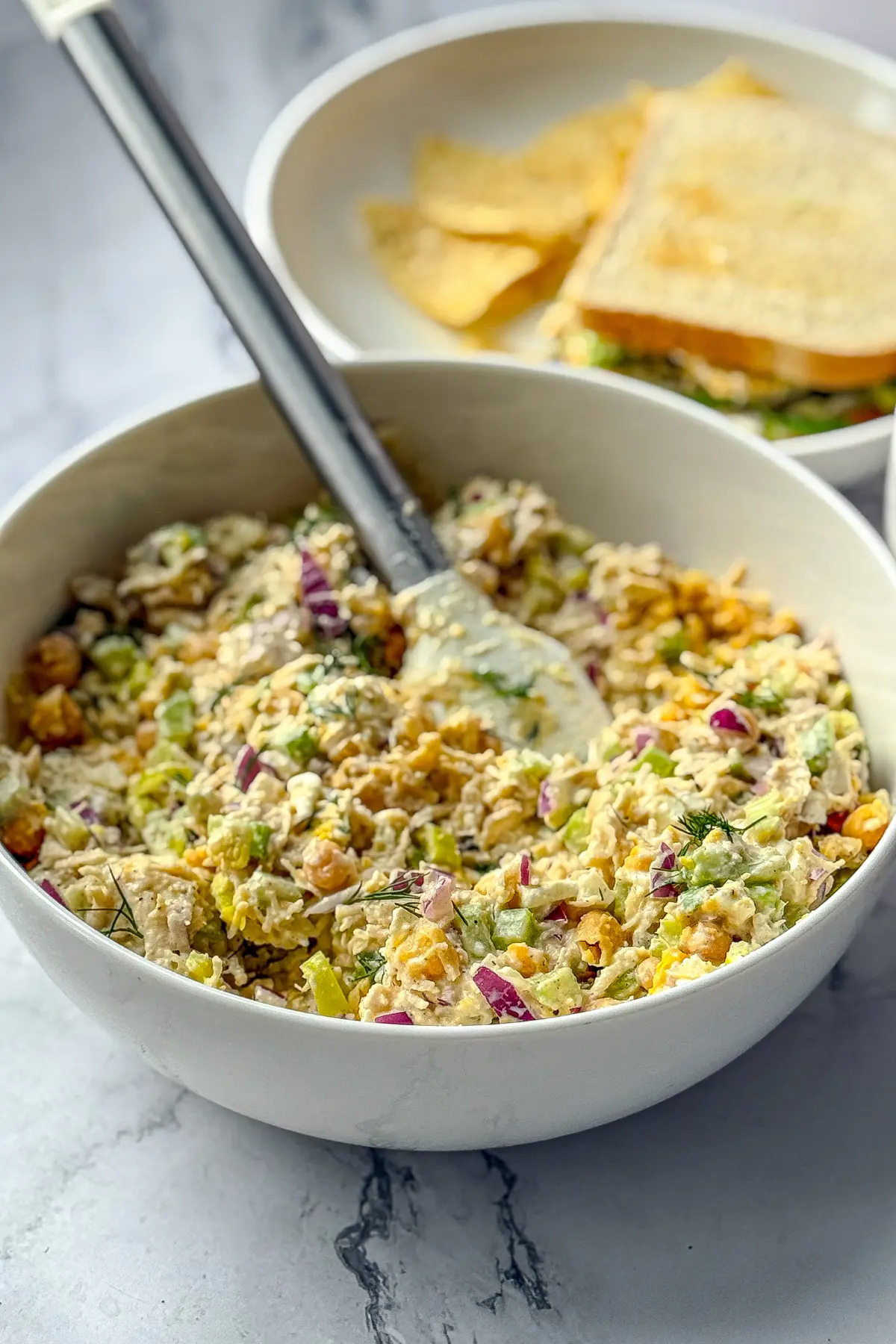 A big bowl of pepperoncini chicken salad with mashed chickpeas with a spatula in it next to a sandwich on a plate.