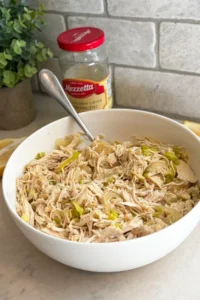 A large white bowl with the shredded and cooked pepperoncini chicken and onions in it with a spoon in it on a counter next to a jar of pepperoncini peppers.
