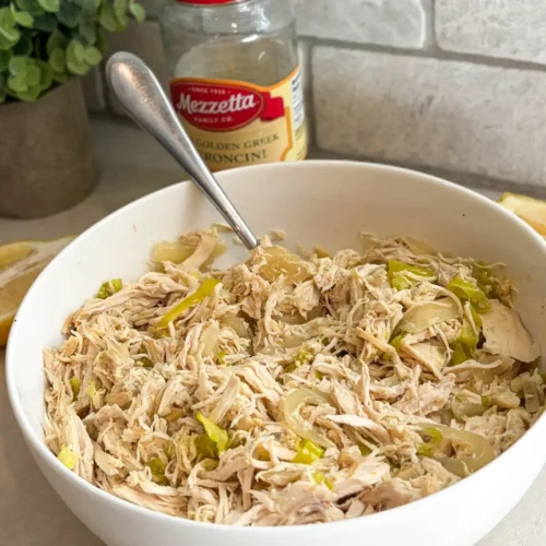 A large white bowl with the shredded and cooked pepperoncini chicken and onions in it with a spoon in it on a counter next to a jar of pepperoncini peppers.