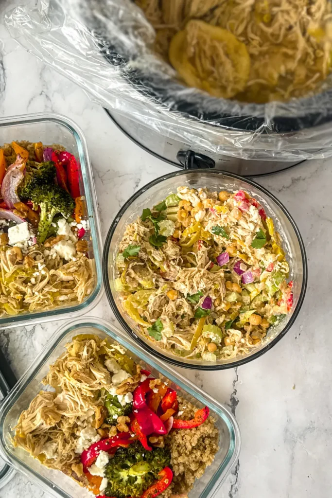 A birds-eye view of a slow cooker with pepperoncini chicken, chicken salad, and meal prep bowls on a counter.