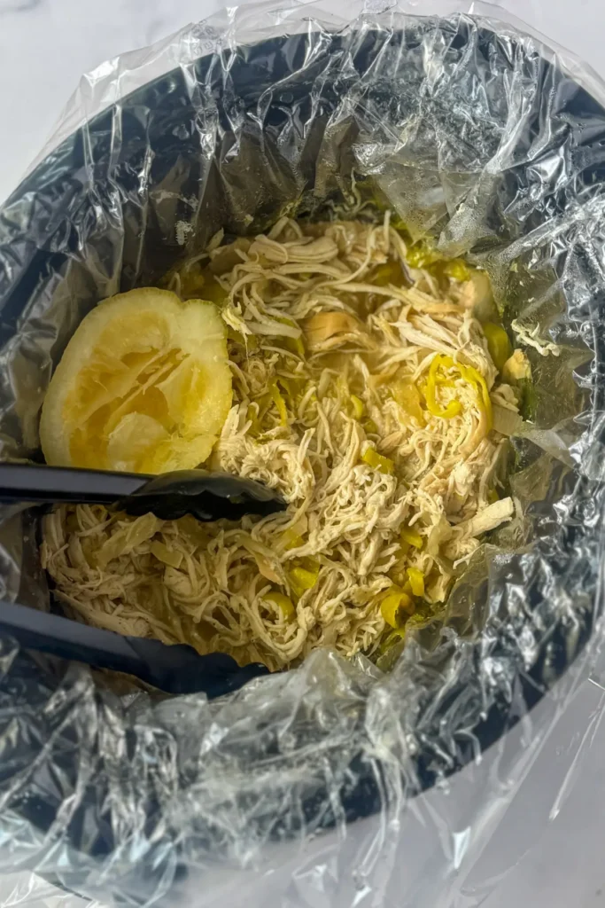 A birds-eye view of the shredded chicken with a lemon wedge in a slow cooker with tongs in it.