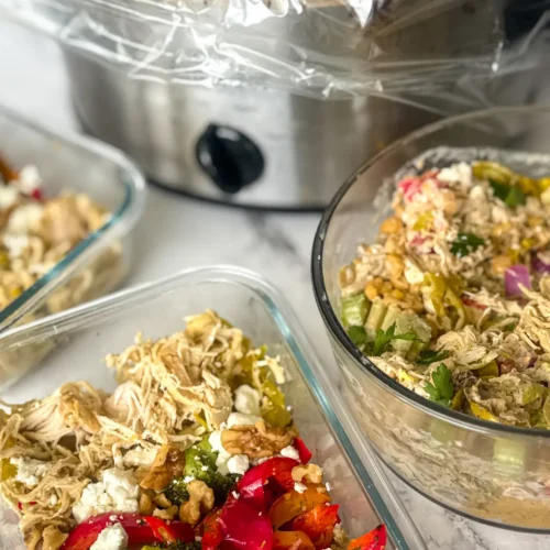 A slow cooker with shredded pepperoncini chicken in it next to a storage contaner of a chicken and quinoa bowl and a big container of chicken salad on a counter.