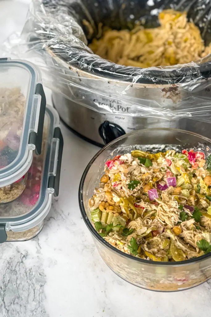 Two meal prep containers stacked on top of each other next to chicken salad and a slow cooker with pepperoncini chicken in it.