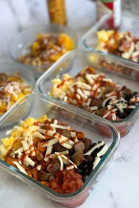 Three savory breakfast bowls next to two sweet mango yogurt bowls on a counter with hot honey and hot sauce.
