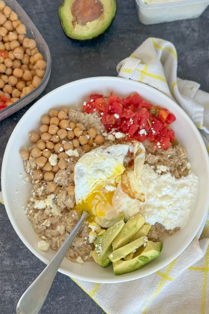 An oatmeal bowl with cottage cheese, eggs, tomatoes, avocado and chickpeas on a table next to an avocado.