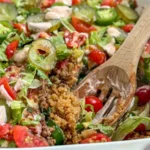 A cheeseburger casserole with hidden lentils topped with lettuce, tomatoes, pickles and yogurt Big Mac sauce in a white baking dish with a wooden spoon in it.