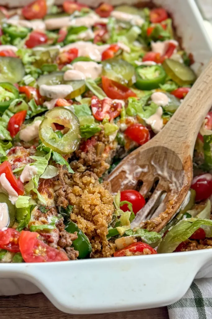 A cheeseburger casserole with hidden lentils topped with lettuce, tomatoes, pickles and yogurt Big Mac sauce in a white baking dish with a wooden spoon in it.