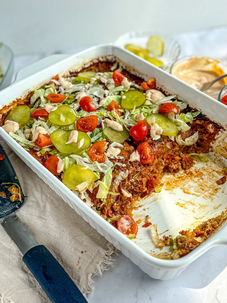 A half eaten casserole topped with shredded lettuce and tomatoes next to a spatula and a bowl of pickles on a counter.