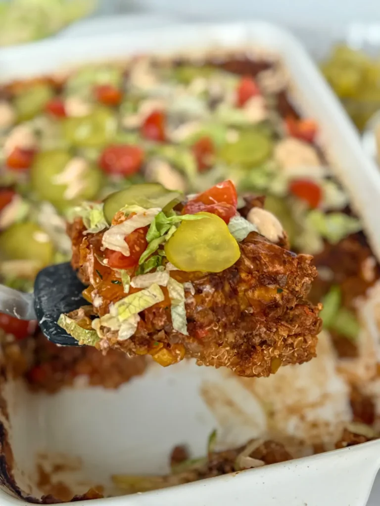A slice of the cheeseburger casserole topped with shredded lettuce and pickles on a spatula over a large white casserole dish.