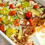 A close-up of a casserole dish with two slices missing of the cheeseburger casserole topped with shredded lettuce, pickle slices and cherry tomatoes.
