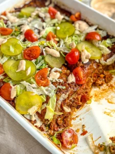 A close-up of a casserole dish with two slices missing of the cheeseburger casserole topped with shredded lettuce, pickle slices and cherry tomatoes.