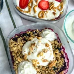A berry fruit cobbler topped with Greek yogurt in a small meal prep container next to a fruit cobbler topped with strawberries and pretzels.
