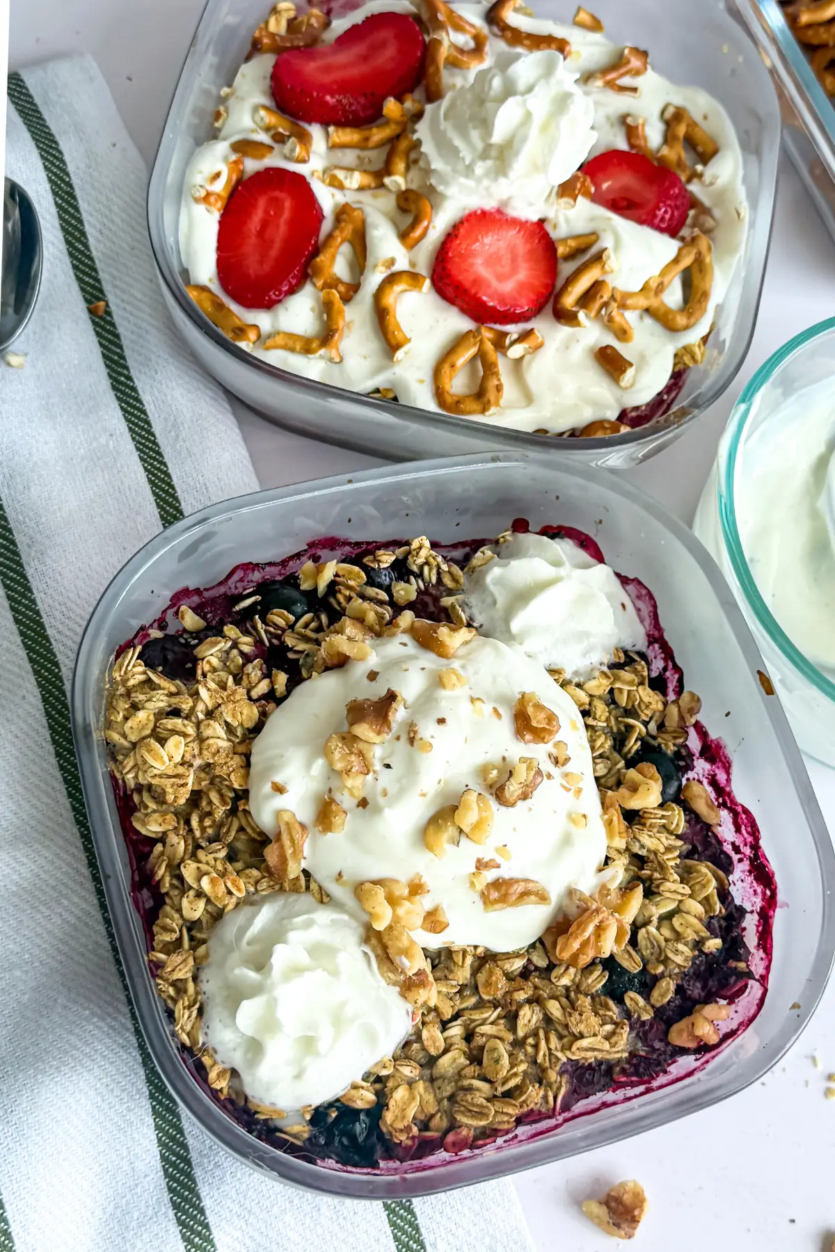 A berry fruit cobbler topped with Greek yogurt in a small meal prep container next to a fruit cobbler topped with strawberries and pretzels.