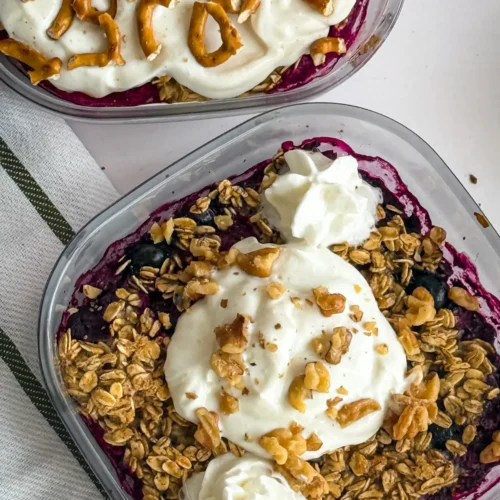 A close-up of the lemon berry cobbler in a meal prep glass container topped with Greek yogurt and whipped cream next to the strawberry pretzel cobbler on a table.
