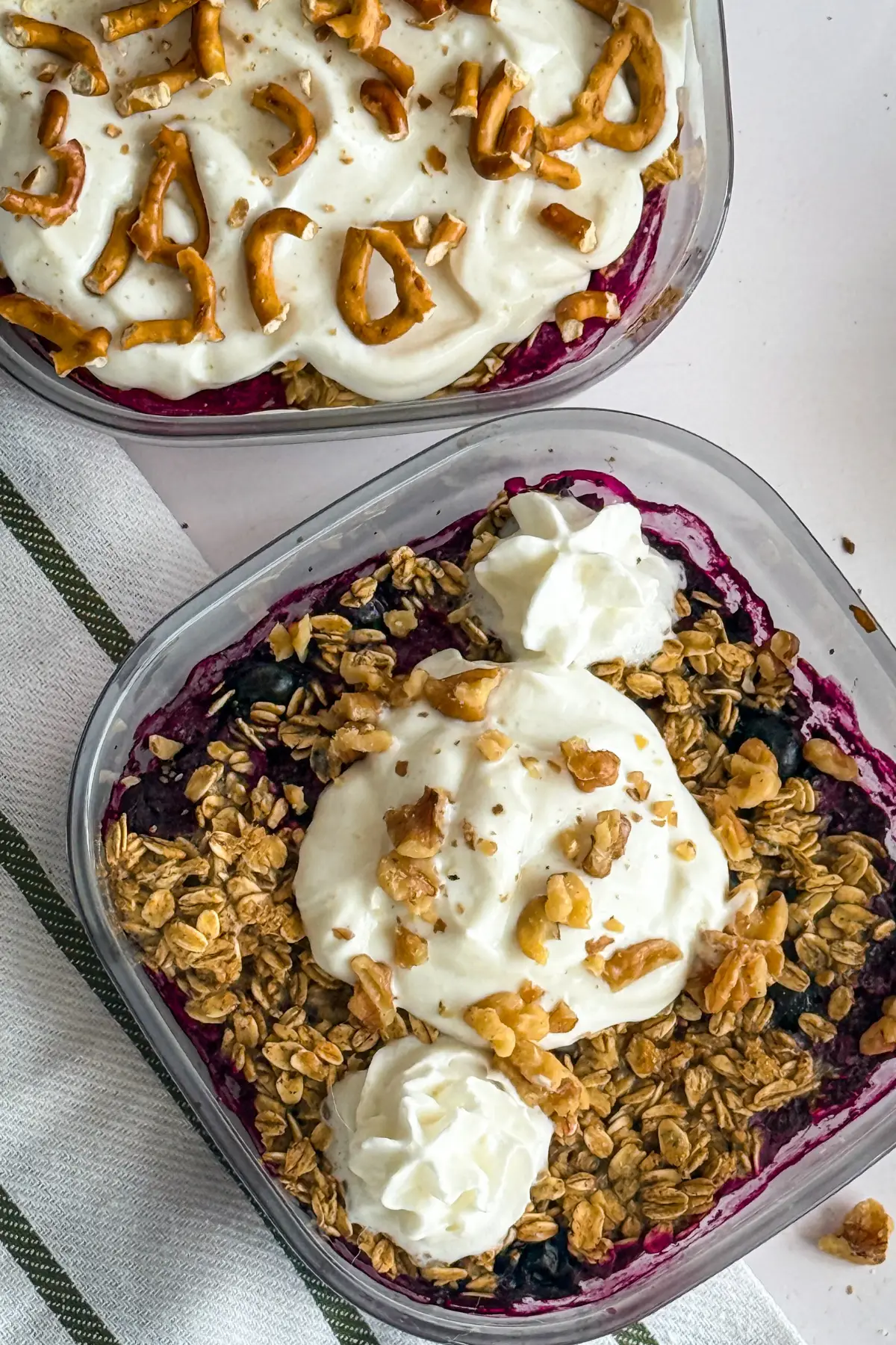 A close-up of the lemon berry cobbler in a meal prep glass container topped with Greek yogurt and whipped cream next to the strawberry pretzel cobbler on a table.