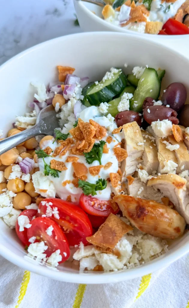 A Greek chicken bowl with olives,fresh veggies, feta cheese and topped with a garlic yogurt in a large bowl on a counter.