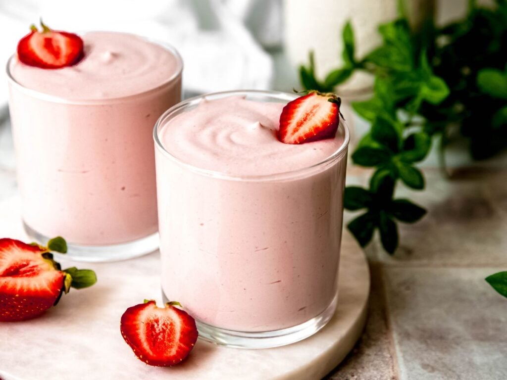 A large glass of strawberry protein fluff garnished with a strawberry on a white platter.