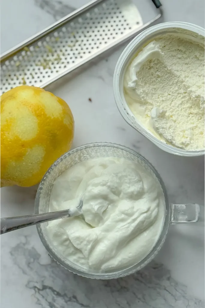 A bowl of yogurt next to a carton of whipped cottage cheese and a lemon with a zester on a counter.