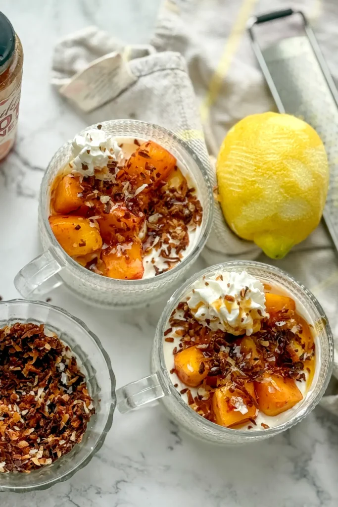 A birds-eye view of two fully loaded and assembled mango cheesecake bowls topped with coconut on a table next to a lemon and a bowl of coconut.
