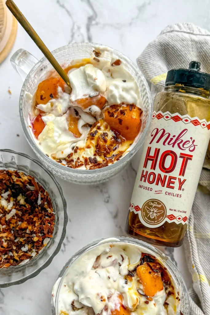 Two bowls of "cheesecake" with toppings mixed into them next to a bottle of hot honey and a bowl of shredded toasted coconut on a counter.