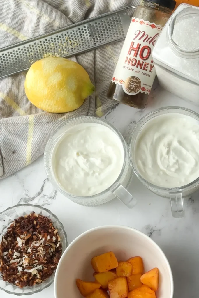 Two yogurt bowls next to mangos, coconuts and hot honey on a counter.