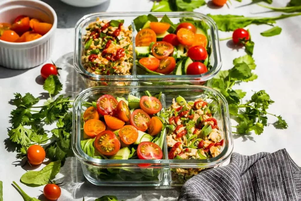 Two meal prep containers with spicy tuna salad and fresh vegetables on a table. 