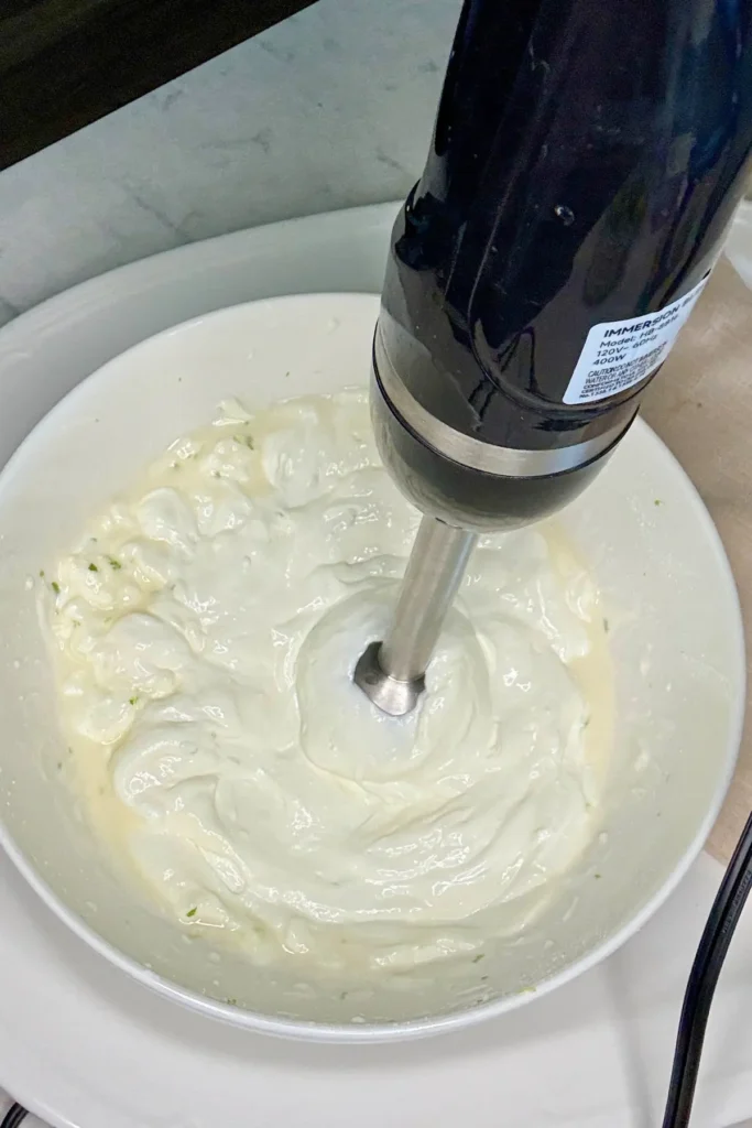 An immersion blender in a large bowl with the key lime pie dip ingredients.