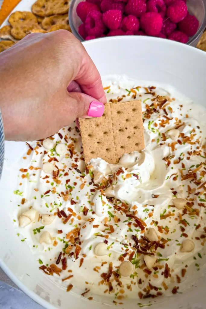 A graham cracker being dipped into a big bowl of key lime pie dip topped with toasted coconut and white chocolate chips.