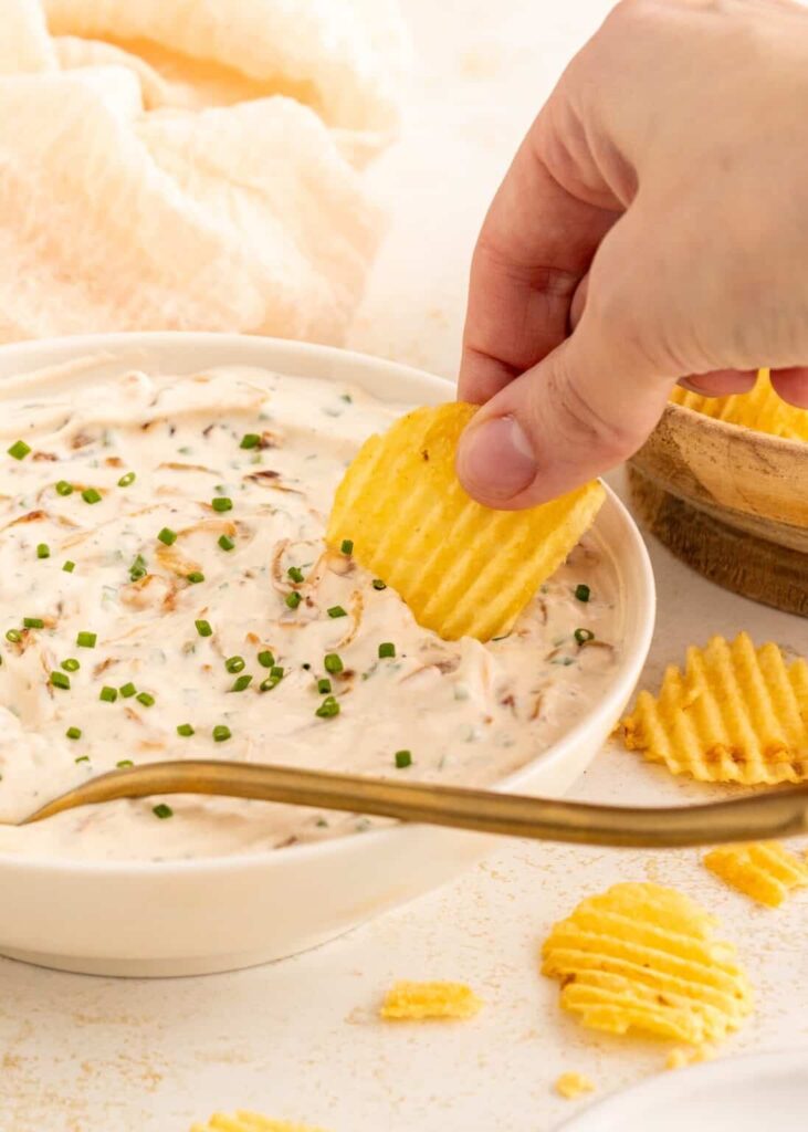A chip digging into a big bowl of French onion dip on a table.