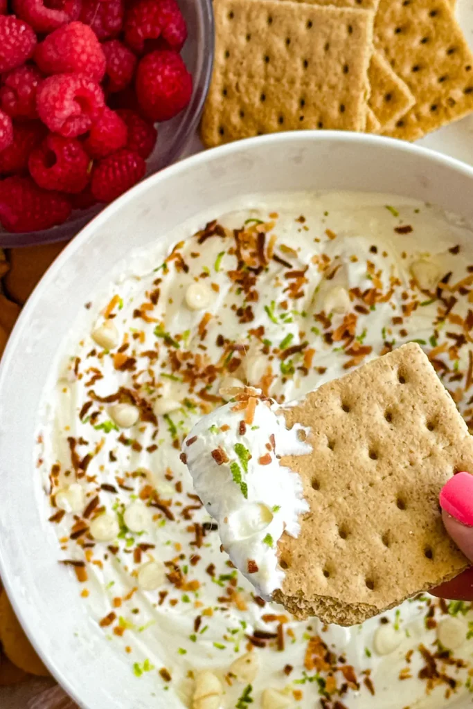 A graham cracker dipped into a big bowl of Greek yogurt dip surrounded by raspberries.